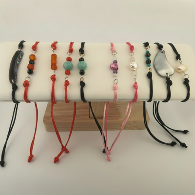 A set of handmade adjustable cord bracelets displayed on a white stand, featuring various bead and shell designs in red, pink, black, orange, and turquoise.